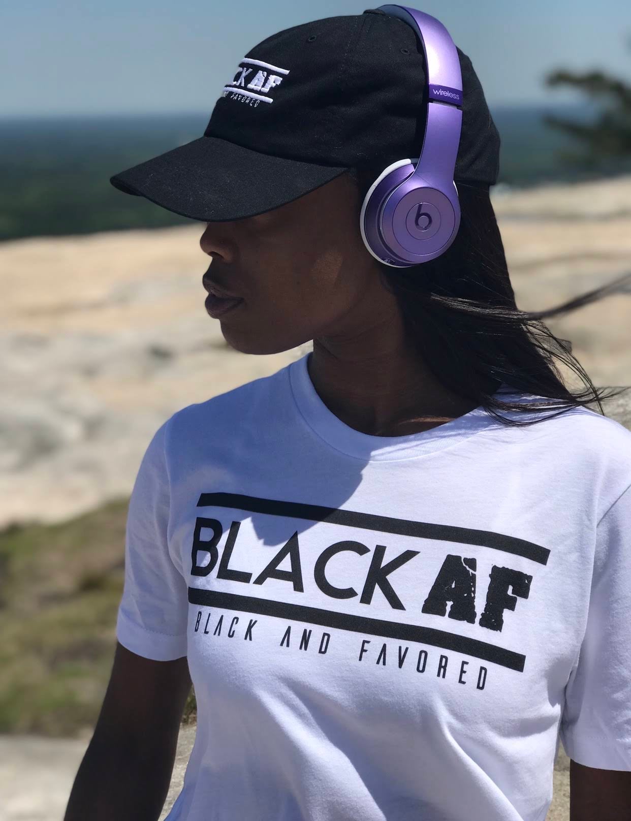 Person wearing a white t-shirt with 'BLACK AF' text, black cap, and purple headphones outdoors.